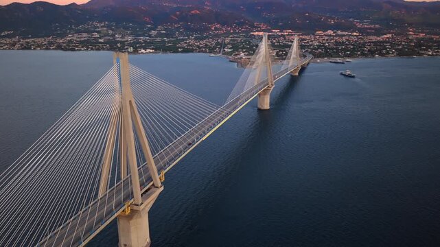 Beautiful aerial perspective of the illuminated Rio-Antirrio bridge in Greece, spanning the gulf of corinth with city lights and mountains visible on the coastline during a colorful sunset