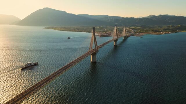 Golden sunset light illuminating the rio antirrio bridge spanning the gulf of corinth in greece, with a ferry boat crossing the calm waters and mountains visible in the background