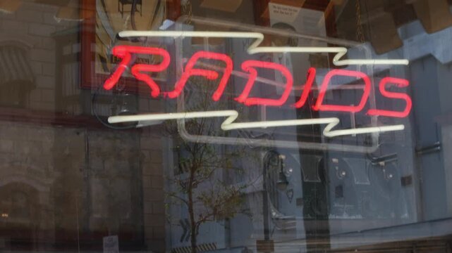 Radios neon sign glowing in a shop window with reflections from nearby buildings. Close-up of vintage storefront lettering and bright red illuminated tubing