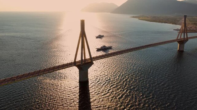 Beautiful aerial perspective of the rio-antirrio bridge in greece with the setting sun reflecting on the corinthian gulf, showing a long shadow and distant mountainous coastline