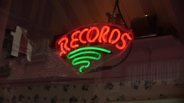 Records neon sign glowing in a storefront window with reflections on the glass. Vintage music shop display featuring bright red lettering and green vinyl record icon