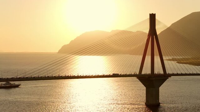 Scenic view of the famous rio antirrio bridge in greece with cars crossing during a beautiful golden sunset, the sun reflecting on the calm waters of the gulf of corinth