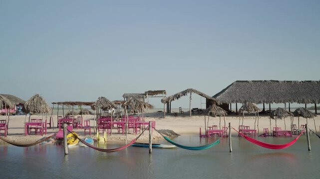 Static wide shot of a tropical beach setup with colorful hammocks over water, palapa huts, and pink outdoor furniture on white sand.
