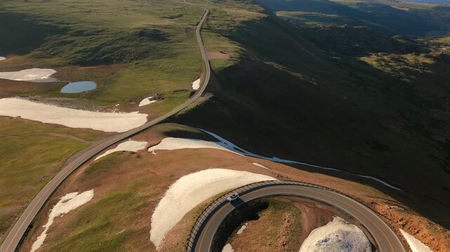 Drone footage captures Beartooth Highway in Montana as vehicles navigate serpentine curves through lush green alpine meadows with melting snow patches and small ponds in golden hour light.