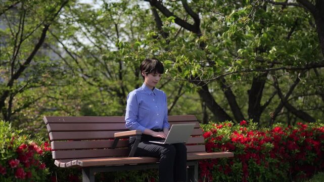 新緑を背景にベンチに座ってノートパソコンのキーボードを打つビジネスウーマンのスローモーションショット