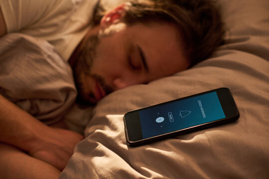 Close up of Caucasian young adult man sleeping beside smartphone alarm on bed, showing morning wake up routine and digital dependence. Ideal for sleep habits, wellness, burnout themes