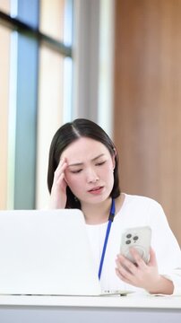 パソコンを前に忙しくて連絡が来るスマホを見たりマルチタスクで悩む考える困るオフィスカジュアルの女性、繁忙期や忙しいタスク管理ビジネスイメージ縦動画　
