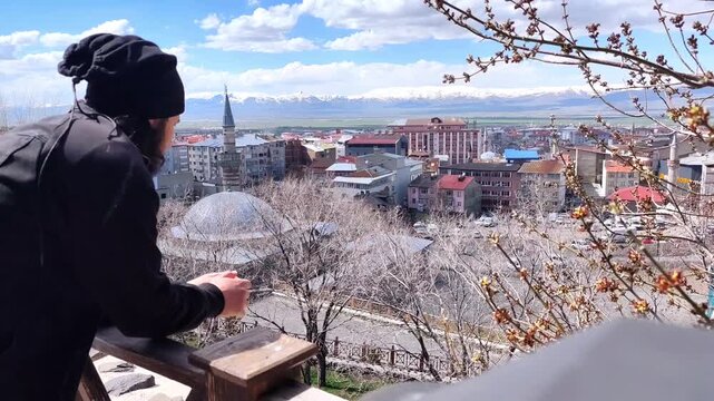 Static side-view shot of a man wearing a black beanie contemplating the historical cityscape of Erzurum with snowy mountains in the distance.