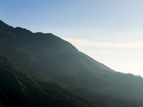 Aerial view of the lush green slopes of Mount Merapi shrouded in light morning mist under a clear blue sky near Kecamatan Pakem, Daerah Istimewa Yogya, Indonesia.