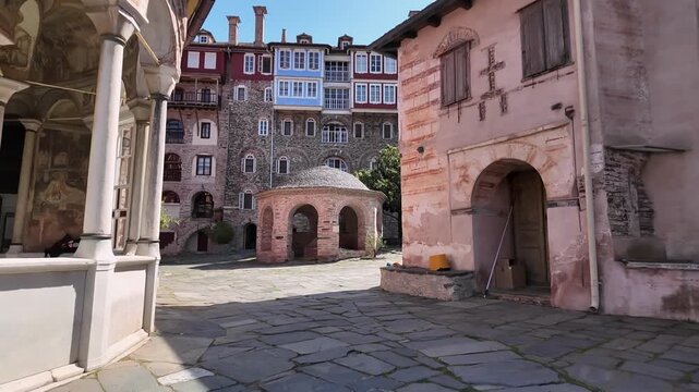 Visitors walk through the courtyard of an old monastery in Mount Athos Greece. The sun lights up the historical stone buildings. Surroundings show a blend of nature and structure.
