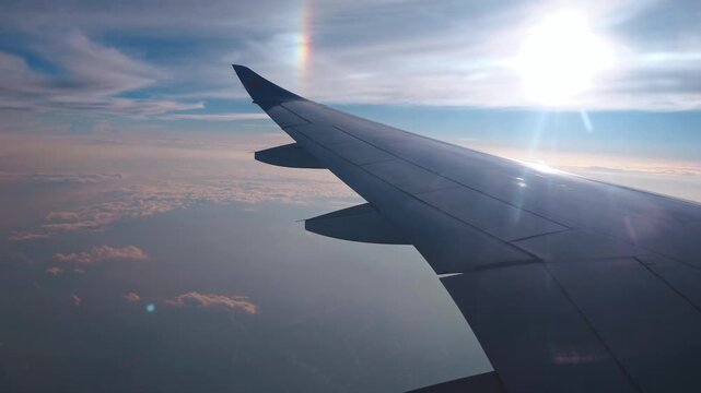 Rare sundog (parhelion) optical phenomenon in the upper atmosphere shot from a passenger window, with aircraft wing is silhouetted against a bright, low-angle sun and a soft, pastel-colored cloud.