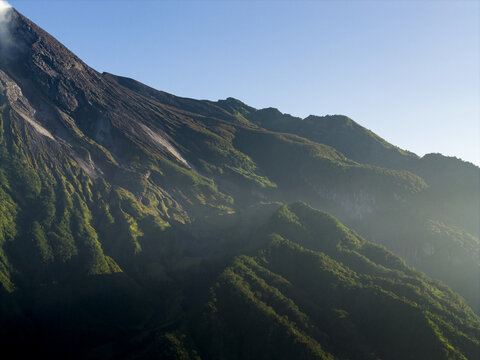 Aerial view of the rugged volcanic slopes of Mount Merapi with lush green vegetation and rocky ridges under a clear blue sky in Kecamatan Pakem, Daerah Istimewa Yogya, Indonesia.
