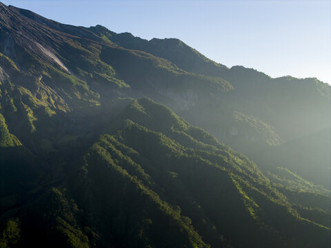 Aerial view of the lush green slopes of Mount Merapi near Bunker Kaliadem with morning sunlight and mist in Kecamatan Pakem, Daerah Istimewa Yogya, Indonesia.