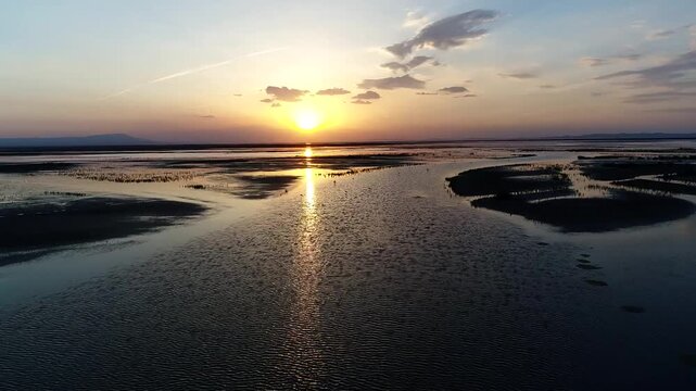 Golden sunset reflects perfectly on calm wetland water with silhouetted reeds and distant birds flying across orange sky in Iranian marsh cinematic aerial stock footage.