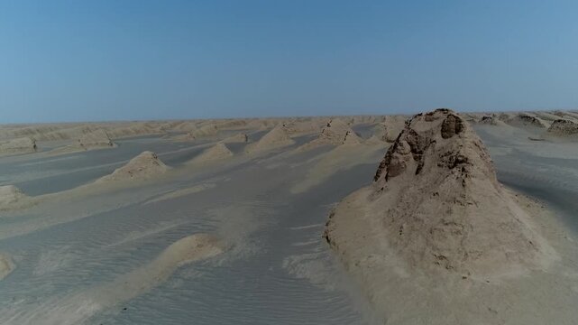 Wind-sculpted rock formation stands alone in vast sandy desert with clear blue sky and distant dunes in central Iran aerial drone cinematic stock footage.