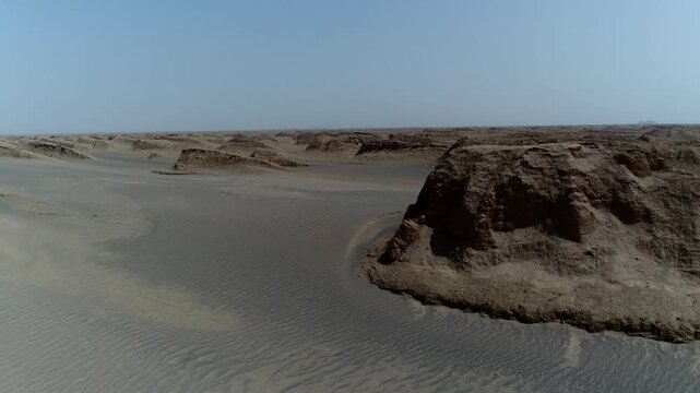 Drone flies low over wind-eroded rock formation with long shadows and surrounding sand dunes in vast Iranian desert under clear blue sky cinematic aerial footage.