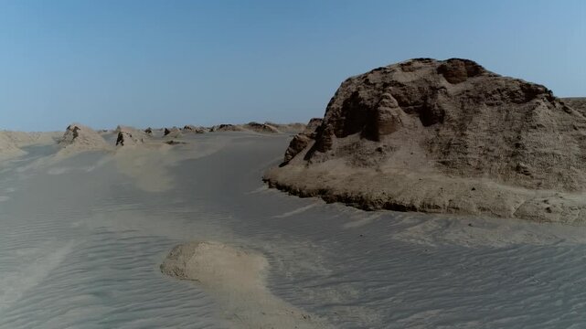 Drone flies over solitary yardang formation with surrounding sand dunes and long shadows under clear sky in Iranian desert cinematic aerial stock video footage.
