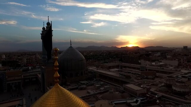 Aerial perspective shows golden dome shining at sunset with historic minarets and pilgrims gathered near the entrance of Lady Masoumeh shrine in Qom Iran cinematic footage.