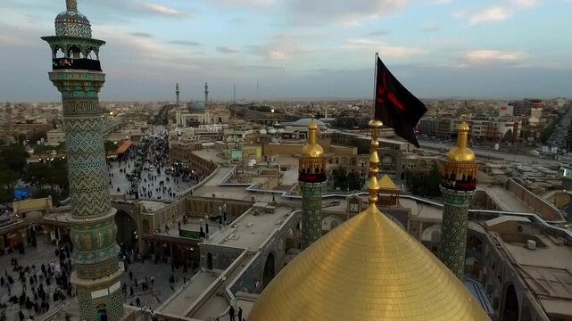 High angle aerial shows golden dome and black flags waving above dense pilgrim crowd in courtyard of Lady Masoumeh shrine at golden hour in Qom Iran stock footage.