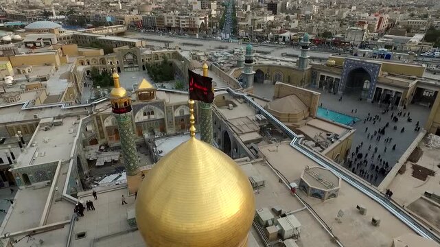 Sunset light highlights golden dome and tall minarets of Lady Masoumeh shrine with black flags and dense crowd of pilgrims in the main courtyard Qom Iran aerial stock video.