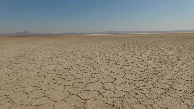 Drone flies low over endless cracked patterns with distant low hills visible on the horizon during harsh bright daylight in central Iranian salt desert stock video production.