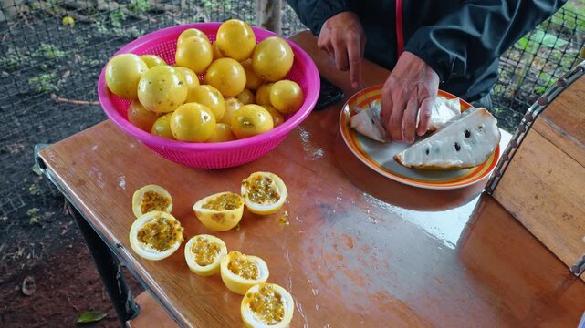 Slicing fresh passion fruit and soursop on table. Hands cutting open fresh yellow passion fruits and a white soursop fruit on a wooden table at a local farm.