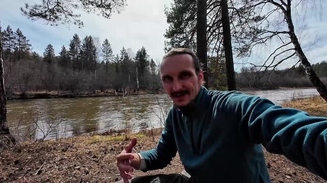 Man camping by river and forest recording selfie vlog on muddy bank pine and birch framing calm water overcast spring sky scattered dry leaves relaxed expression subtle sunlight peaceful