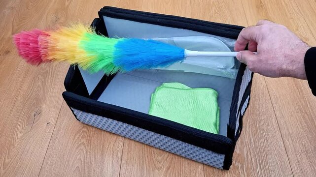 Hand holds colorful feather duster above gray storage box with green cloth on wooden floor, highlighting household cleaning, organization, and practical everyday utility