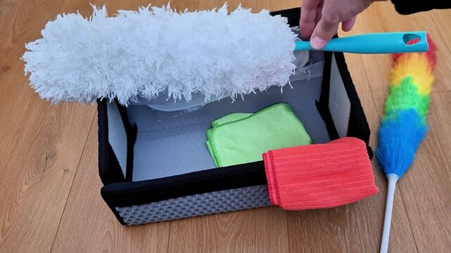 Cleaning kit on wood floor. Hand places white fluffy duster into organizer box with green and red microfiber cloths. Static duster nearby. Practical household chores and organization