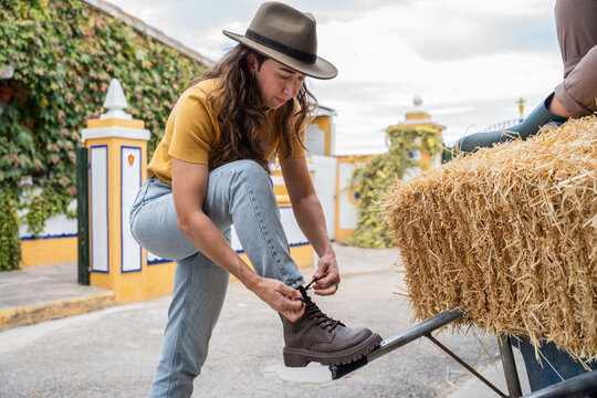Woman tying boot lace beside hay bale on rural farmyard