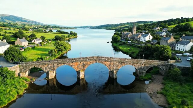 Aerial view of a picturesque stone bridge over a river in a charming countryside village surrounded by rolling green hills.