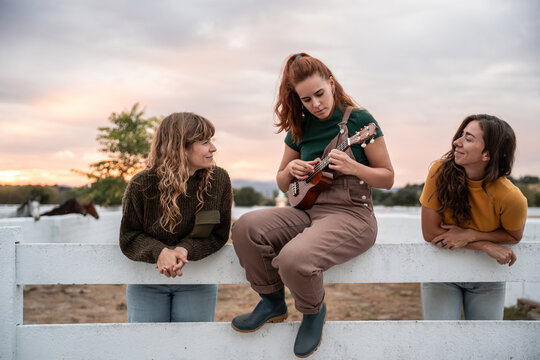 Three women on a fence at sunset listening to ukulele