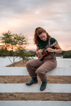 Young woman playing ukulele on fence at sunset outdoors