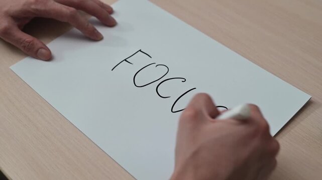 A hand writes the word focus on a sheet of paper. The writing style transitions from lowercase to uppercase, with an emphasis on correct spelling and accentuation