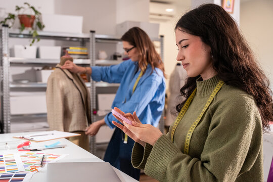Multiethnic women in atelier measuring garment and texting