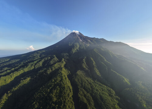 Aerial view of the Mount Merapi volcano with smoke rising from the crater above lush green forested slopes under a clear blue sky near Kecamatan Pakem, Daerah Istimewa Yogya, Indonesia.