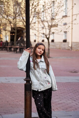 Fototapeta premium A young girl stands playfully next to a vintage pole, showcasing her stylish outfit. Surrounded by an inviting plaza, she captivates the moment with joy under the soft daylight.