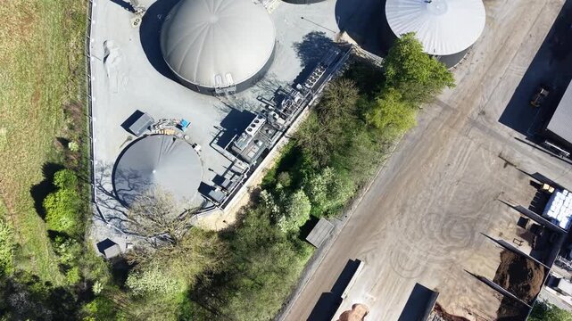 Vertical drone perspective moving over large circular anaerobic digester tanks and outdoor biomass storage bays at a Danish renewable energy site.