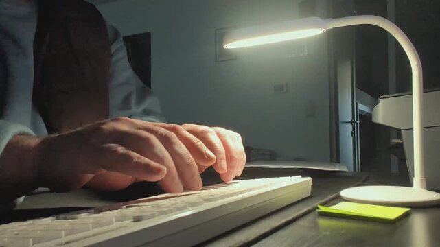 Mans hands typing on a computer keyboard at a desk in a room with a lamp in the evening hours