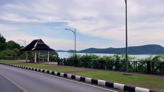 A moving view from the road to the Khao Khad View Point on Cape Panwa in Phuket. A long walkway lined with pavilions overlooks the sea, hills, and tropical coastline of Thailand.