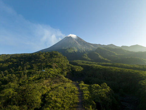 Aerial view of Bunker Kaliadem Merapi with lush green forests and a smoking volcanic peak under a clear blue sky in Kecamatan Pakem, Daerah Istimewa Yogya, Indonesia.