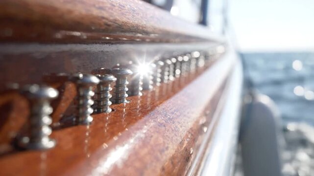 Polished wooden boat rail with gleaming metal screws in shallow focus, sunlit texture and sparkling sea in background conveying warm nautical calm