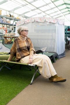Mature woman wearing glasses and poncho, sitting on a cot in a camping supply store