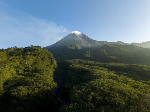 Aerial view of the majestic Mount Merapi volcano with steam rising from its crater above a dense tropical forest under a clear blue sky in Kecamatan Pakem, Daerah Istimewa Yogya, Indonesia.