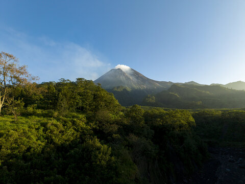 Aerial view of Mount Merapi volcano with smoke rising from the crater above lush green forests under a clear blue sky in Kecamatan Pakem, Daerah Istimewa Yogya, Indonesia.