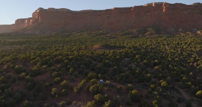 Desert juniper woodland landscape beneath dramatic red mesa cliffs near Kanab Utah at sunset, aerial panoramic ascend