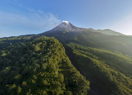 Aerial view of Mount Merapi volcano with smoke rising from its crater above the lush green forests of the Kaliadem area in Kecamatan Pakem, Daerah Istimewa Yogya, Indonesia.