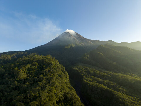 Aerial view of Mount Merapi volcano with smoke rising from the crater above lush green forests under a clear blue sky in Kecamatan Pakem, Daerah Istimewa Yogya, Indonesia.