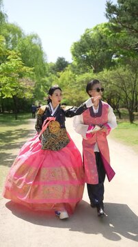 Two women walk side by side through a park. One wears a bright pink skirt with gold patterns. The other wears a matching top with black trousers. They both smile, appear happy and relaxed