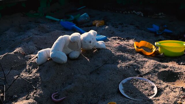Forgotten white stuffed bunny toy and colorful plastic toys lying abandoned in a sandbox, symbolizing lost childhood, loneliness, and the passage of time with shadows slowly moving across the sand